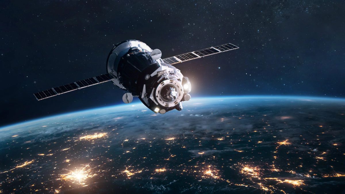 Spacecraft with extended solar panel arrays orbiting above Earth at night, with glowing city lights visible on the surface below.